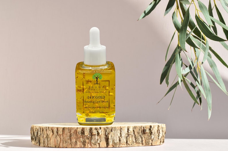 Puede incluir: Una botella de vidrio transparente con un l&iacute;quido dorado y escamas doradas en su interior. La botella tiene un gotero blanco y una etiqueta que dice "24K GOLD Botanical Face Serum". La botella est&aacute; sobre una superficie de madera.