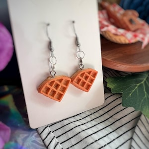May include: A pair of orange waffle earrings with silver-colored hooks. The earrings are displayed on a white card against a background of a striped fabric and other decorative items.
