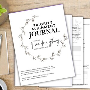 May include: A white "Priority Alignment Journal" with a floral wreath design and the text "Because I can do anything but not everything." The journal is open to several pages, with a spiral-bound notebook, pen, and coffee cup nearby.