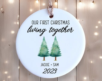May include: White round ceramic ornament with the text "Our First Christmas living together" and two green watercolor trees with gold lights. Below the trees is the text "Jackie + Sam 2023".