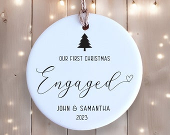 May include: White round ceramic ornament with a black silhouette of a Christmas tree and the text "Our First Christmas Engaged" with a small heart symbol. Below the text is "John & Samantha 2023".