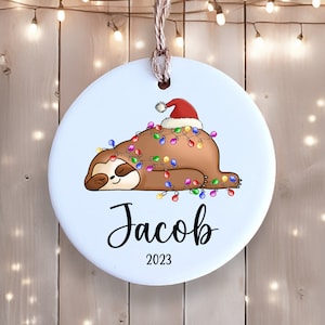 May include: A white ceramic Christmas ornament featuring a cartoon sloth wearing a Santa hat and tangled in colorful Christmas lights. The ornament is personalized with the name "Jacob" and the year "2023".