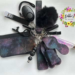May include: A collection of black accessories, including a personal alarm, a pom-pom keychain, a small zippered pouch, and a lip gloss. The items are connected by keyrings. The pouch and keychains have a spiderweb design.