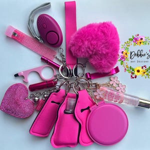 Op de afbeelding: Een verzameling roze accessoires aan sleutelhangers, waaronder een persoonlijk alarm, een pluizig hart, een lipgloss en een flessenhouder. De items zijn gerangschikt rond een centrale sleutelhanger.