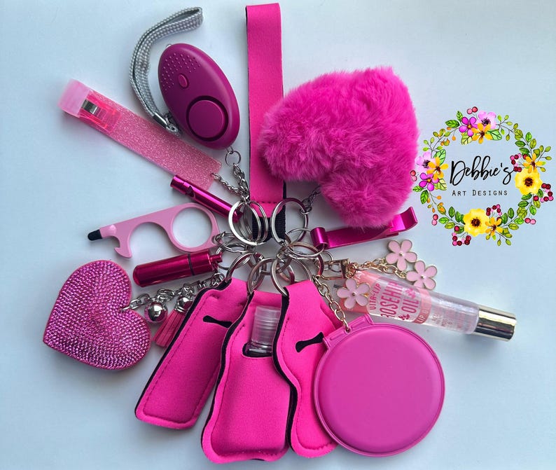 Op de afbeelding: Een verzameling roze accessoires aan sleutelhangers, waaronder een persoonlijk alarm, een pluizig hart, een lipgloss en een flessenhouder. De items zijn gerangschikt rond een centrale sleutelhanger.