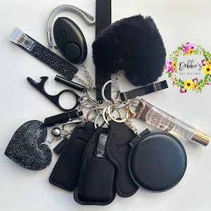 May include: A collection of black accessories on a white background, including a keychain with a fluffy heart, a personal alarm, a lip gloss, and a bottle holder. The keychain also includes a bottle opener and a nail clipper.