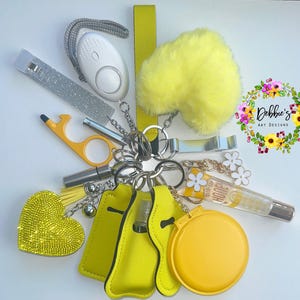 May include: A collection of yellow accessories on a white surface, including a fluffy heart-shaped keychain, a personal safety alarm, a nail file, and a lip gloss. The items are connected by silver keychains.