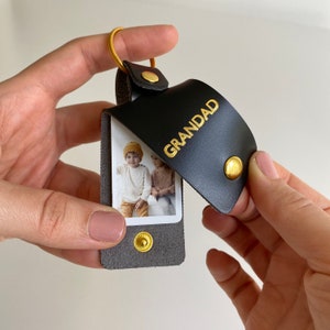 May include: A black leather keychain with a gold ring and a photo insert. The photo insert shows two children. The keychain has the word "GRANDAD" embossed in gold letters.