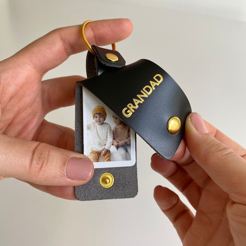 May include: A black leather keychain with a gold ring and a photo insert. The photo insert shows two children. The keychain has the word "GRANDAD" embossed in gold letters.