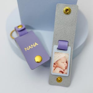 May include: Two personalized keychains. One is lavender with "NANA" in gold lettering and a gold snap. The other is gray with a photo of a baby and a lavender loop and gold snaps. Both are on a light blue surface.
