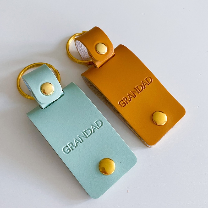 May include: Two leather keychains with gold rings and metal accents. Both keychains are rectangular and have the word "GRANDAD" embossed on them. One keychain is light blue and the other is brown.