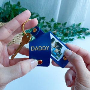 May include: A blue leather keychain with a photo insert and the word "DADDY" embossed in gold. The keychain has a gold key ring and a small gold button.