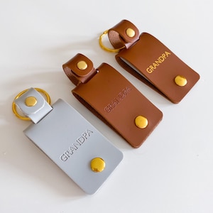 May include: Three leather keychains with gold key rings and metal accents. Each keychain has the word "GRANDPA" embossed on the front in gold lettering. One keychain is gray, and the other two are brown.