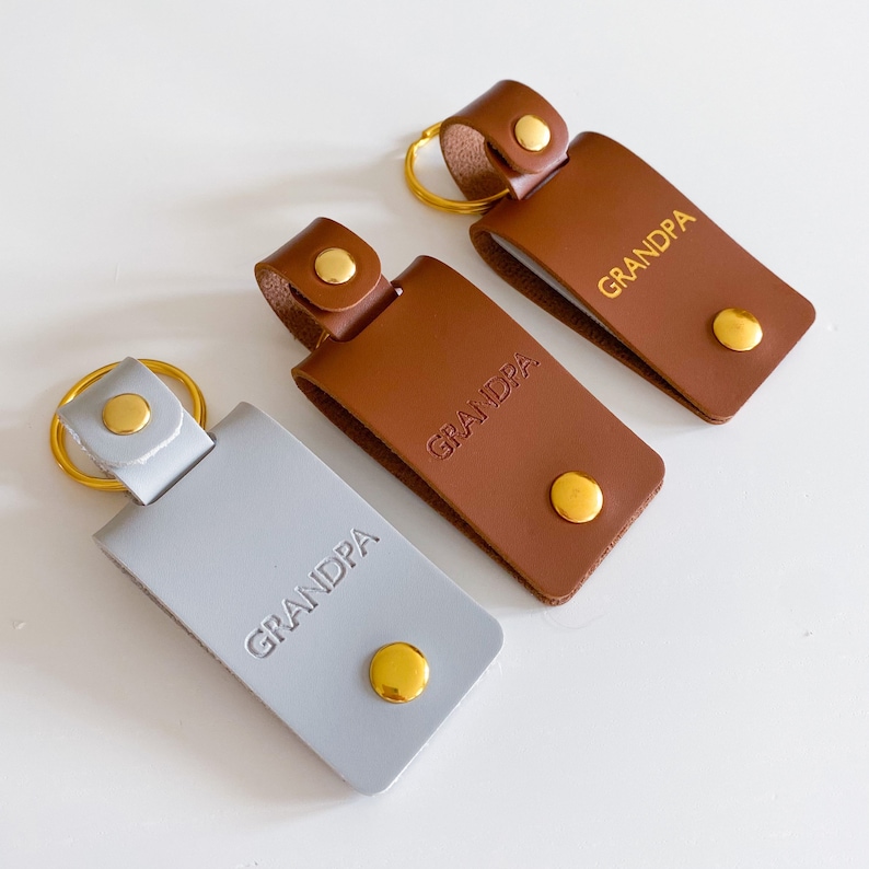 May include: Three leather keychains with gold key rings and metal accents. Each keychain has the word "GRANDPA" embossed on the front in gold lettering. One keychain is gray, and the other two are brown.