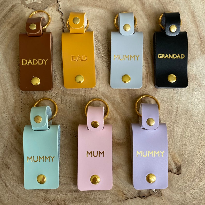 May include: Six leather keychains with gold rings and gold lettering. The keychains are brown, yellow, gray, black, light blue, and lavender. The lettering on the keychains reads "Daddy", "Dad", "Mummy", "Grandad", "Mummy", and "Mum".