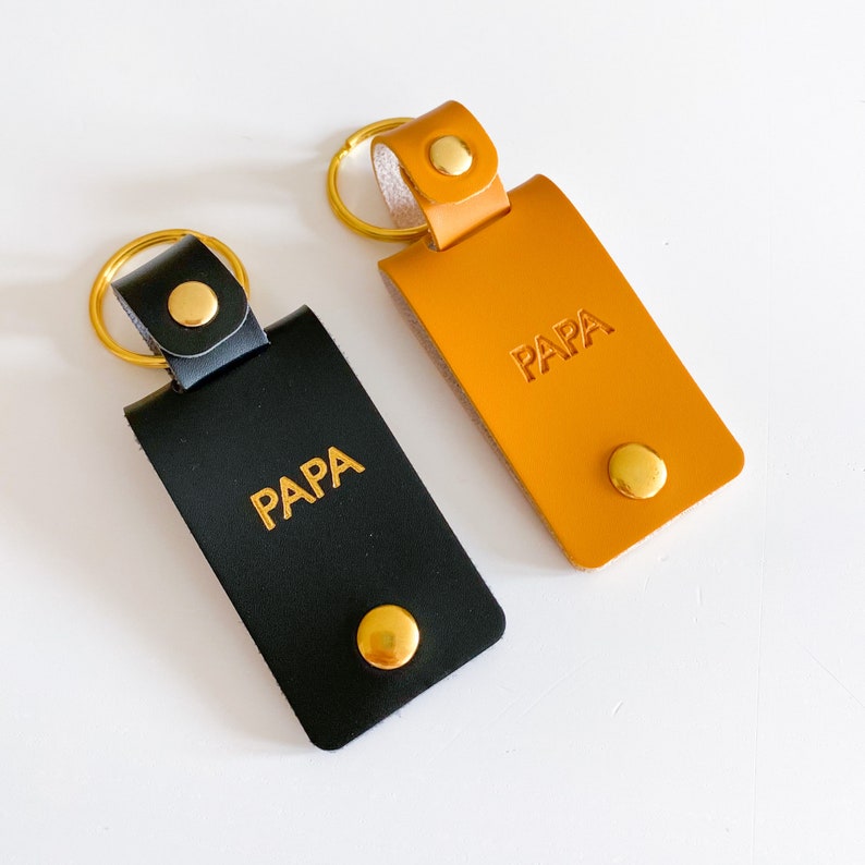 May include: Two leather keychains with gold key rings and gold metal accents. Both keychains are rectangular and have the word "PAPA" embossed in gold lettering. One keychain is black leather and the other is orange leather.