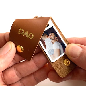 May include: A brown leather keychain with a photo insert. The photo shows a father and son hugging. The keychain is embossed with the word "DAD".