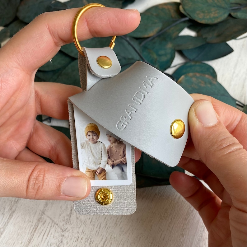 May include: A gray leather keychain with a photo insert featuring two children. The keychain has a gold ring and a gold snap closure. The leather flap has the word "GRANDMA" embossed on it.