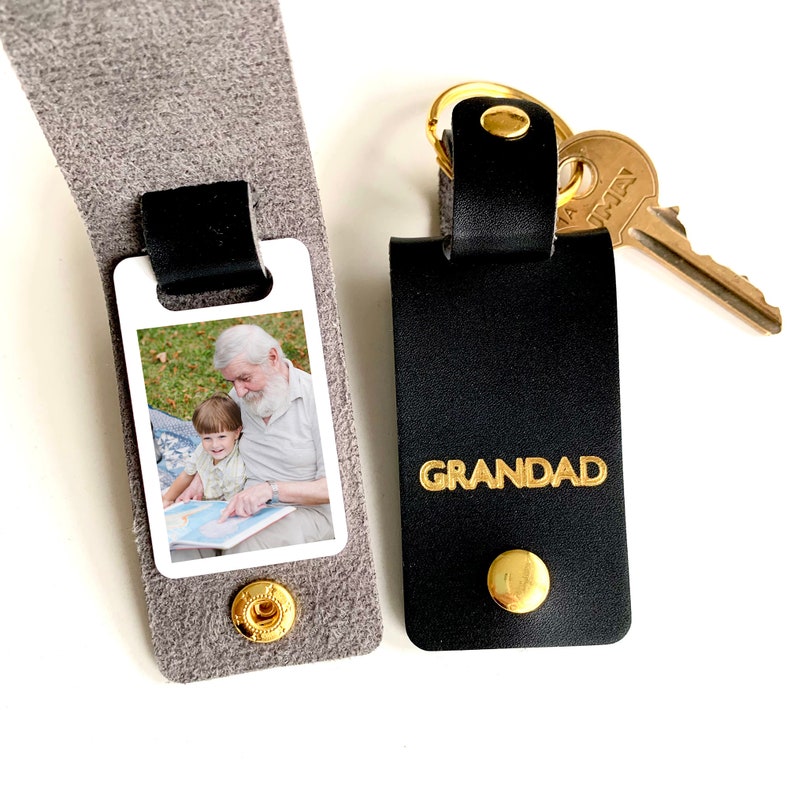 May include: A black leather keychain with a gold key ring and a photo insert. The photo insert shows a man and a child reading a book. The keychain has the word "GRANDAD" embossed in gold letters.