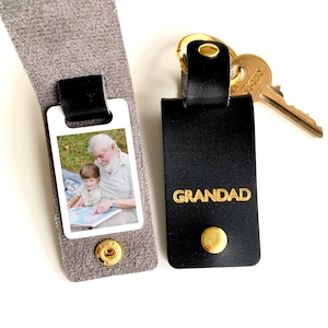 May include: A black leather keychain with a gold key ring and a photo insert. The photo insert shows a man and a child reading a book. The keychain has the word "GRANDAD" embossed in gold letters.