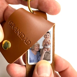 May include: A brown leather keychain with the word "GRANDAD" embossed on the front. The keychain has a small photo insert that shows a family of three: a grandfather, grandmother, and a young boy.