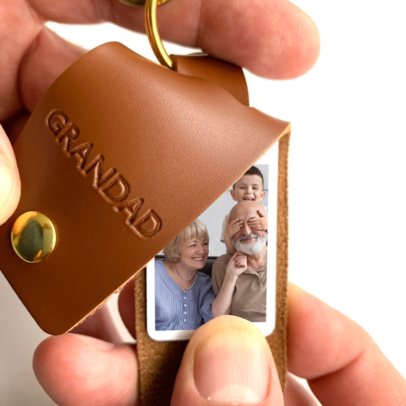 May include: A brown leather keychain with the word "GRANDAD" embossed on the front. The keychain has a small photo insert that shows a family of three: a grandfather, grandmother, and a young boy.