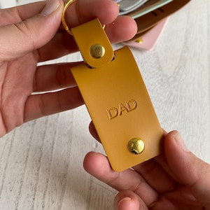 May include: A mustard yellow leather keychain with a gold ring and two gold rivets. The keychain has the word "DAD" embossed on it.