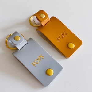 May include: Two leather keychains with gold rings and gold metal accents. Both keychains have the word "POPS" embossed in gold lettering. One keychain is gray and the other is orange.