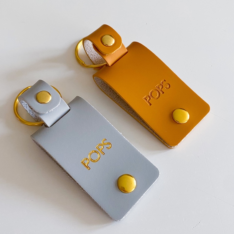 May include: Two leather keychains with gold rings and gold metal accents. Both keychains have the word "POPS" embossed in gold lettering. One keychain is gray and the other is orange.