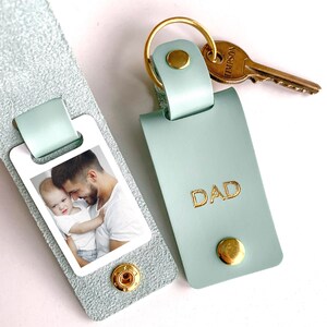 May include: A light green leather keychain with a photo of a father and son. The keychain has a gold metal ring and a small gold metal tag that says "DAD".
