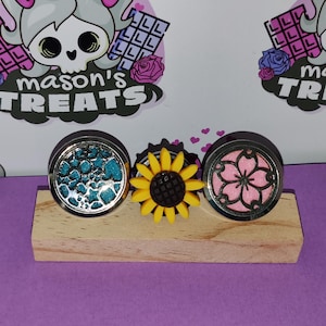 May include: Three decorative plugs on a small wooden block. One has a teal and silver design, the second is a yellow sunflower, and the third features a pink cherry blossom design. The background includes the text "mason's TREATS" and a cartoon skull.