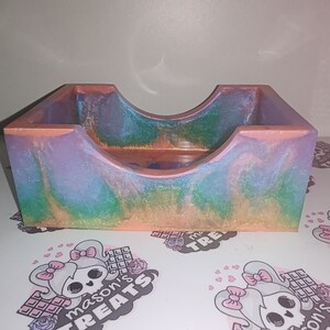 May include: A small, iridescent, resin trinket dish with a rainbow swirl pattern. The dish is made of clear resin with a light pink, blue, and green swirl pattern. The dish has a rounded, U-shaped opening and a flat base.