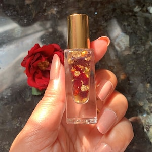 May include: A clear glass bottle with a gold cap filled with a red liquid and gold flakes. The bottle is being held in a hand.