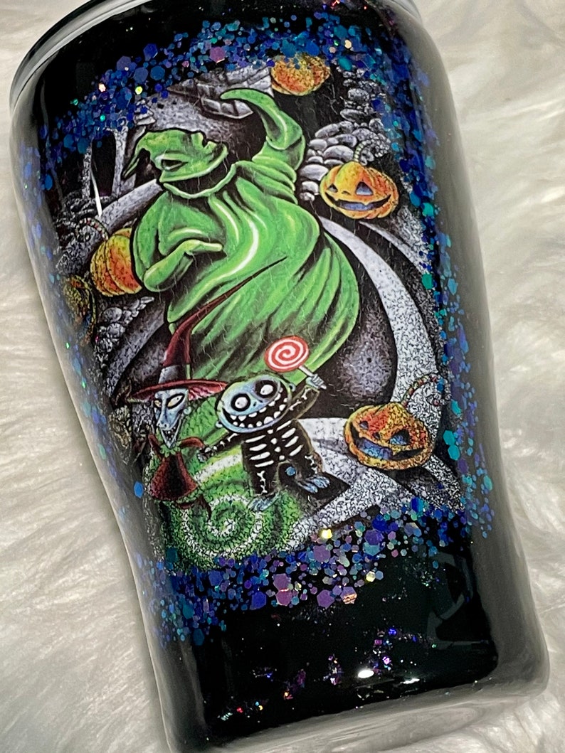 May include: A black tumbler with a glittery blue finish features a colourful illustration of characters from The Nightmare Before Christmas. The characters include Jack Skellington, Sally, and Oogie Boogie.