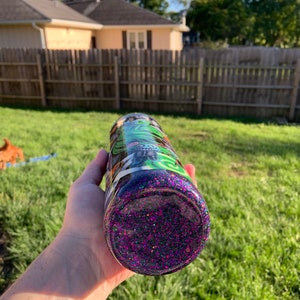 May include: A purple glitter tumbler with a black and green design featuring a skull and crossbones. The tumbler is made of stainless steel and has a double-walled construction to keep drinks cold or hot.