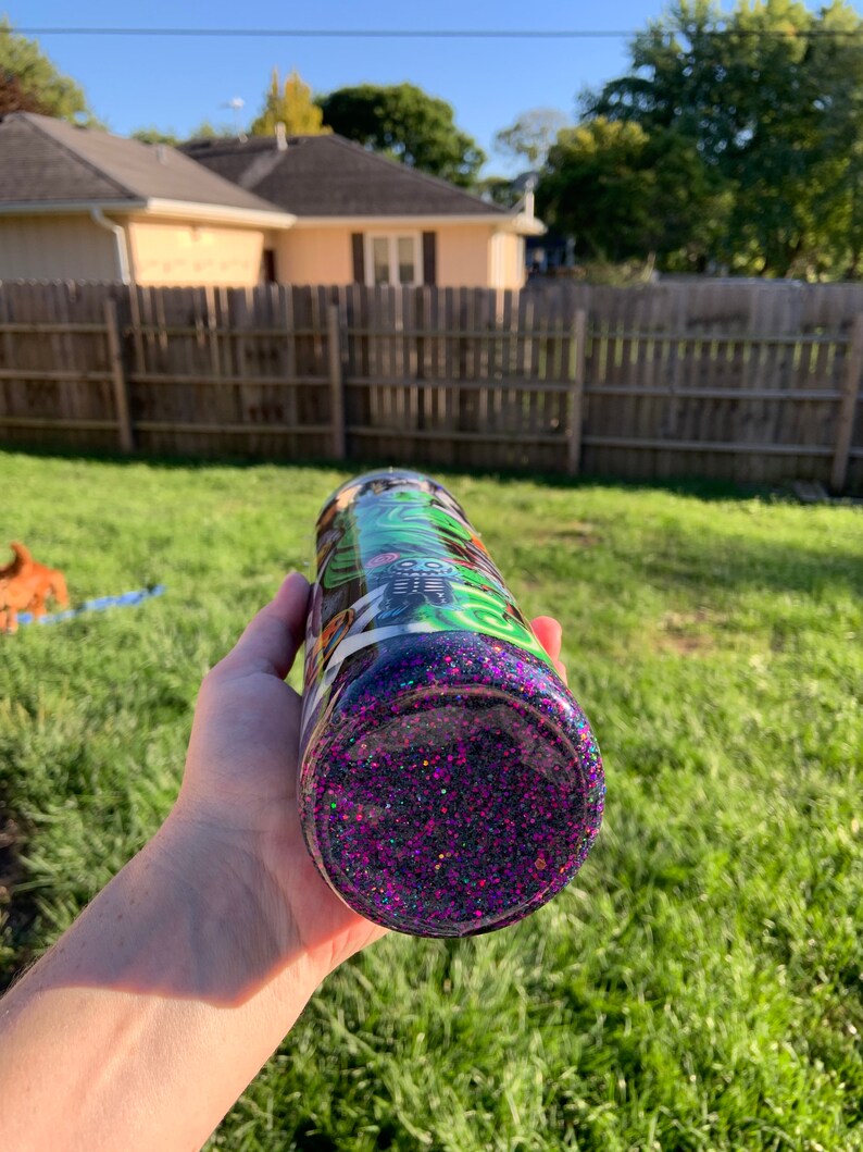 May include: A purple glitter tumbler with a black and green design featuring a skull and crossbones. The tumbler is made of stainless steel and has a double-walled construction to keep drinks cold or hot.