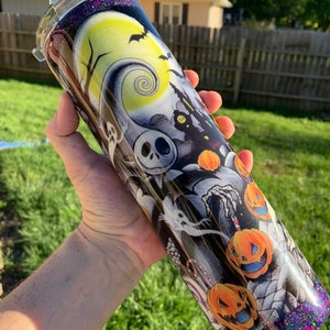 May include: A black and white tumbler with a glittery purple bottom. The tumbler features a Halloween-themed design with Jack Skellington, pumpkins, and a spooky moon.