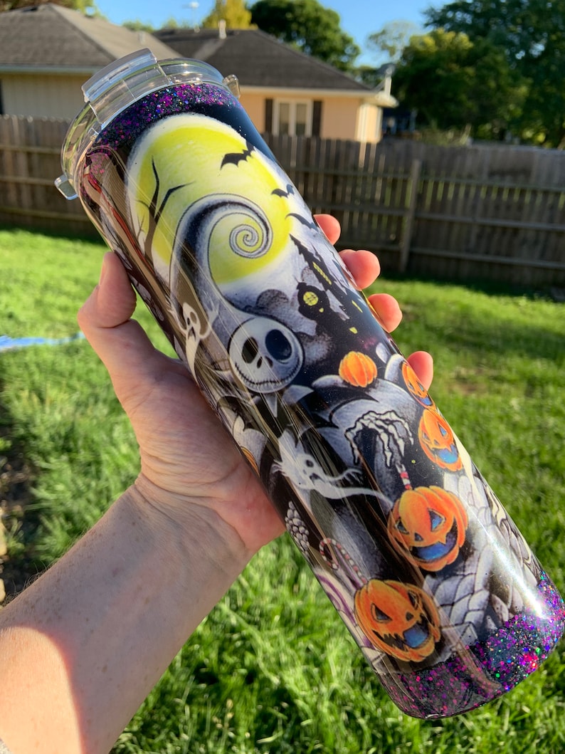 May include: A black and white tumbler with a glittery purple bottom. The tumbler features a Halloween-themed design with Jack Skellington, pumpkins, and a spooky moon.