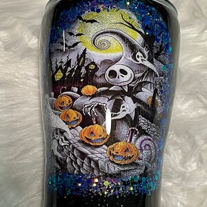 May include: A black and white tumbler with a glittery blue and green design. The tumbler features a scene from the Nightmare Before Christmas movie with Jack Skellington and pumpkins.