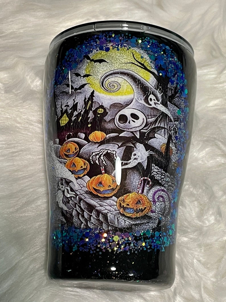 May include: A black and white tumbler with a glittery blue and green design. The tumbler features a scene from the Nightmare Before Christmas movie with Jack Skellington and pumpkins.