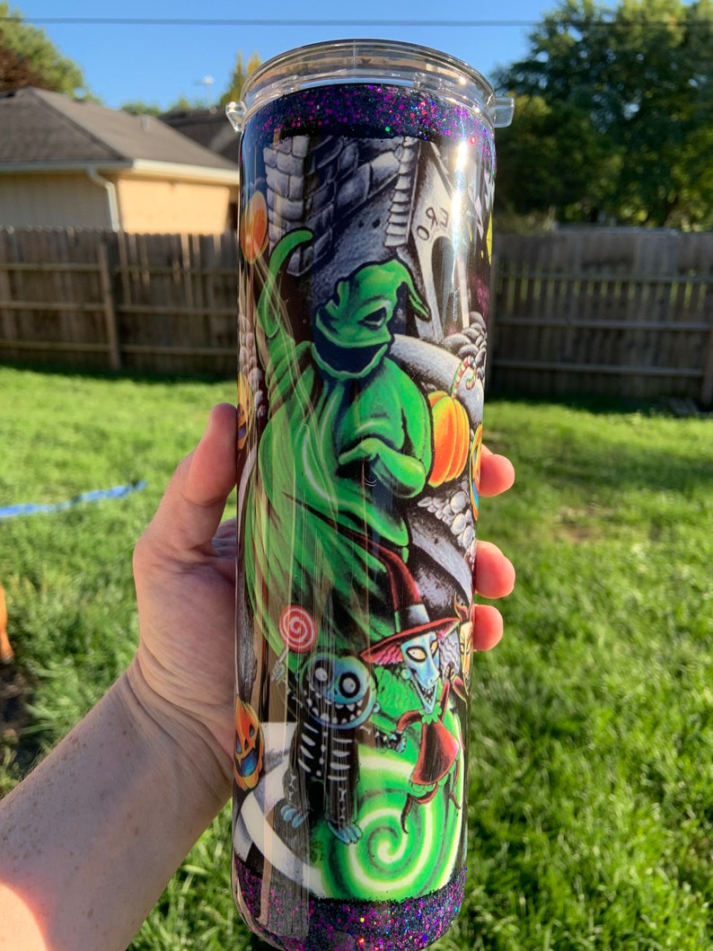 May include: A black and green tumbler with a glitter finish. The tumbler features a graphic of characters from the Nightmare Before Christmas, including Jack Skellington, Sally, and Oogie Boogie.