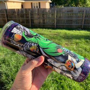 May include: A black and green stainless steel tumbler with a Halloween-themed design featuring a ghost, pumpkins, and candy. The tumbler has a purple glitter finish.