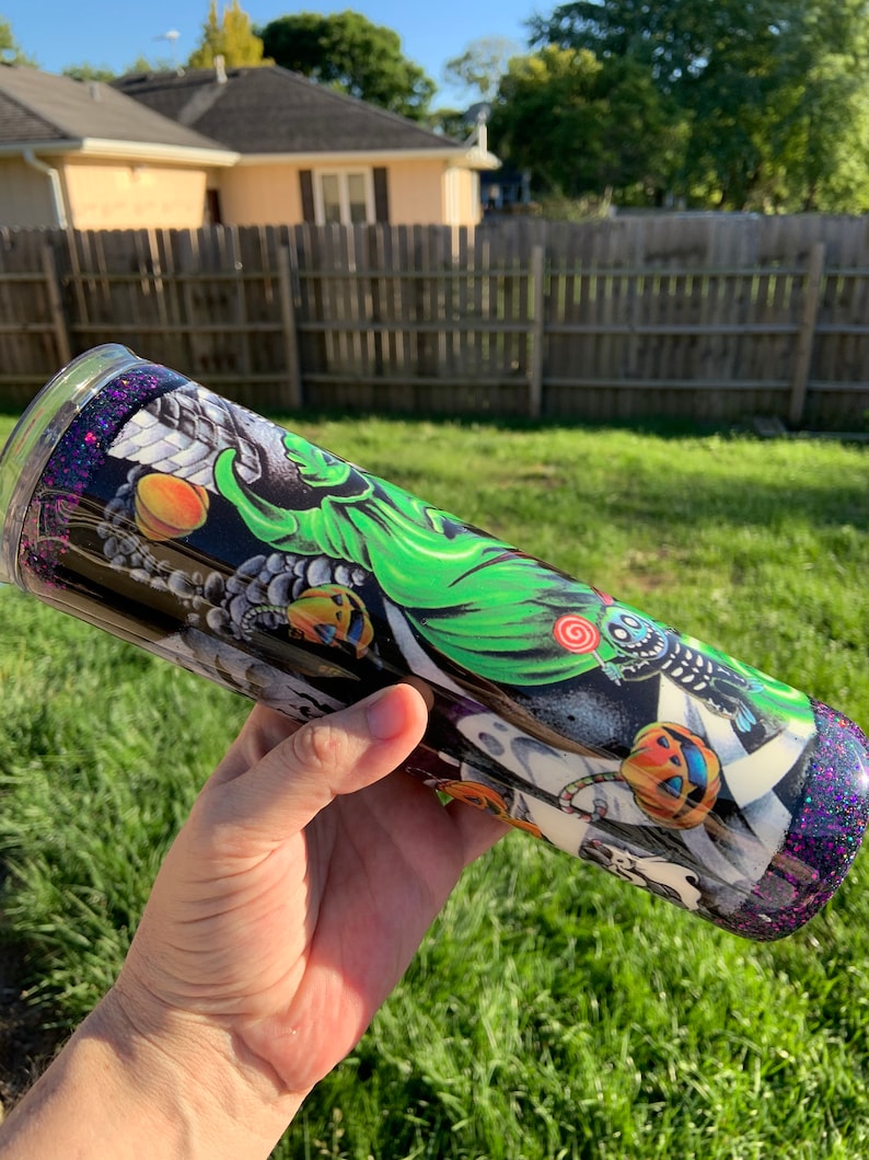May include: A black and green stainless steel tumbler with a Halloween-themed design featuring a ghost, pumpkins, and candy. The tumbler has a purple glitter finish.
