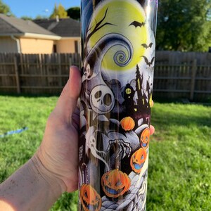 May include: A black and white tumbler with a glittery purple bottom. The tumbler features a Halloween-themed design with a spooky tree, a full moon, a haunted house, and jack-o'-lanterns.