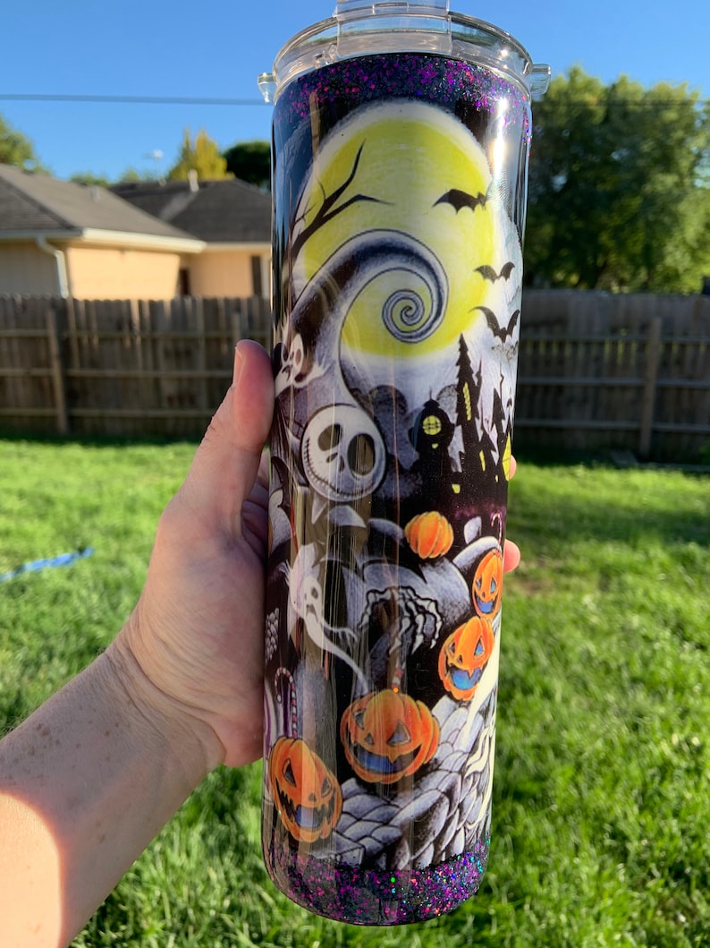 May include: A black and white tumbler with a glittery purple bottom. The tumbler features a Halloween-themed design with a spooky tree, a full moon, a haunted house, and jack-o'-lanterns.