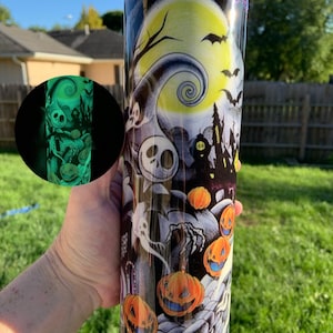 May include: A black and white tumbler with a glow-in-the-dark design featuring a spooky Halloween scene with pumpkins, ghosts, and a jack-o-lantern. The design includes a crescent moon with a spiral pattern and bats flying in the night sky.