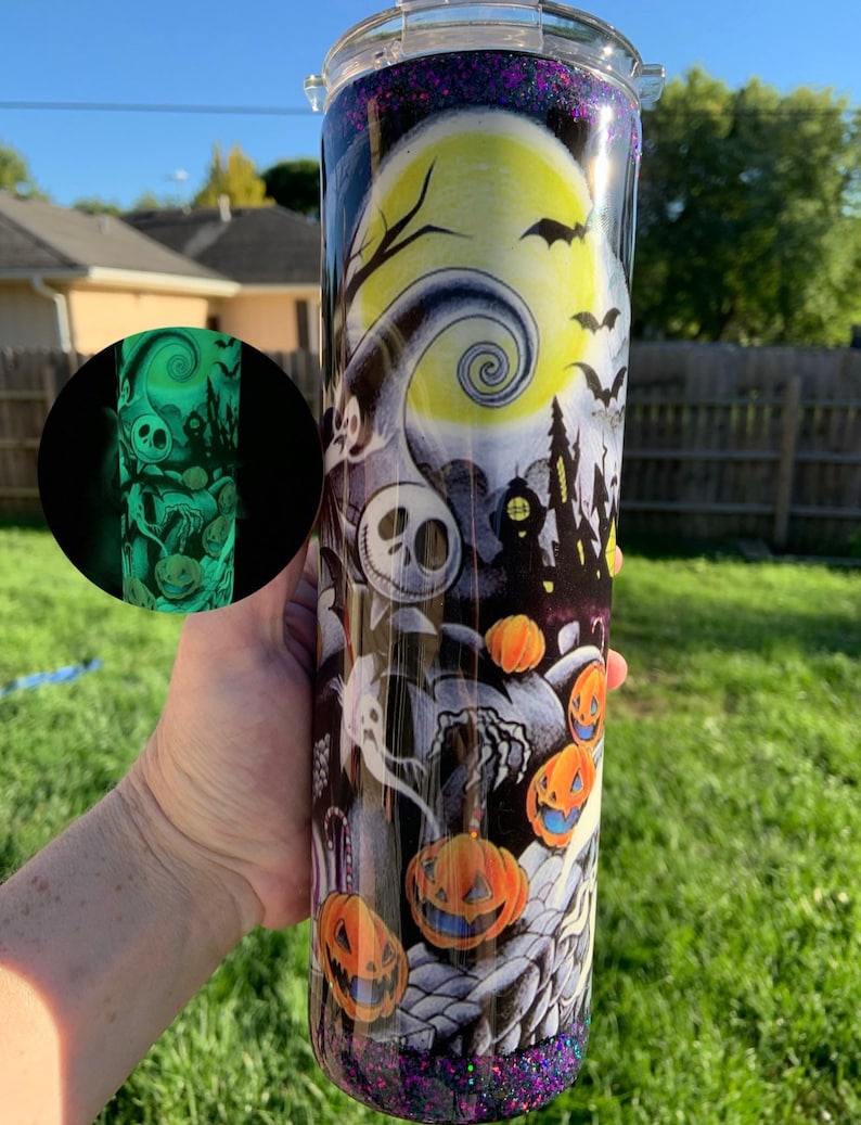 May include: A black and white tumbler with a glow-in-the-dark design featuring a spooky Halloween scene with pumpkins, ghosts, and a jack-o-lantern. The design includes a crescent moon with a spiral pattern and bats flying in the night sky.