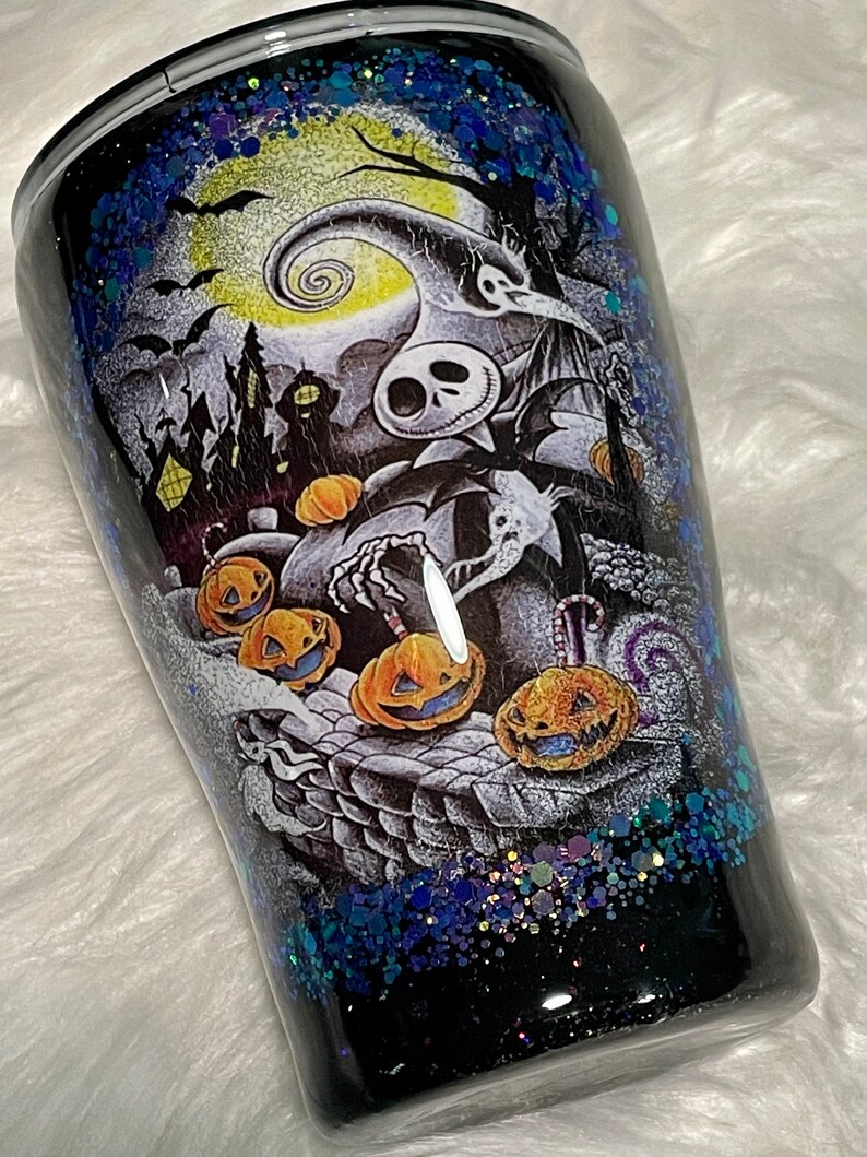 May include: A black and white tumbler with a glittery blue and purple finish. The tumbler features a graphic of Jack Skellington from The Nightmare Before Christmas with pumpkins and a spooky Halloween scene.