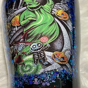 May include: A black tumbler with a glittery blue finish features a colourful illustration of characters from The Nightmare Before Christmas. The characters include Jack Skellington, Sally, and Oogie Boogie.