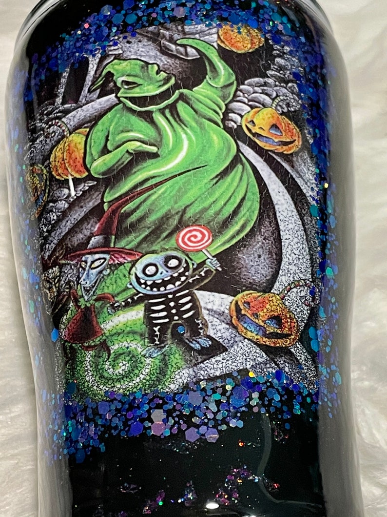 May include: A black tumbler with a glittery blue finish features a colourful illustration of characters from The Nightmare Before Christmas. The characters include Jack Skellington, Sally, and Oogie Boogie.